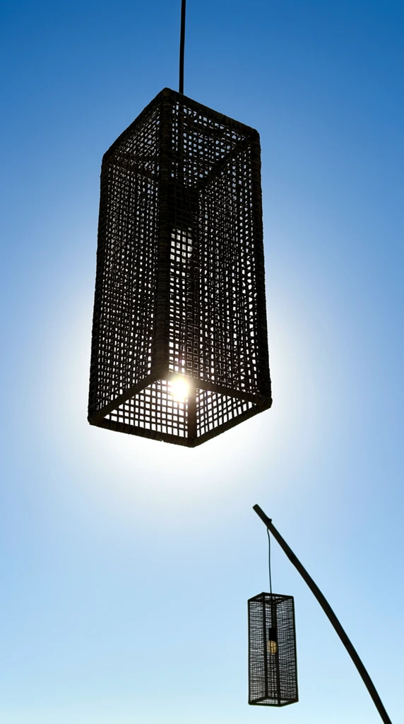 rectangular woven pendant lamp hanging against a bright blue sky, dark rattan cage structure with fine square grid pattern, silhouette backlit by intense sunlight directly behind it, sun flare piercing through the bottom edge mesh, low angle perspective looking upward, second smaller matching lantern suspended from an angled pole in the lower right, minimalist composition, high contrast, clear sky