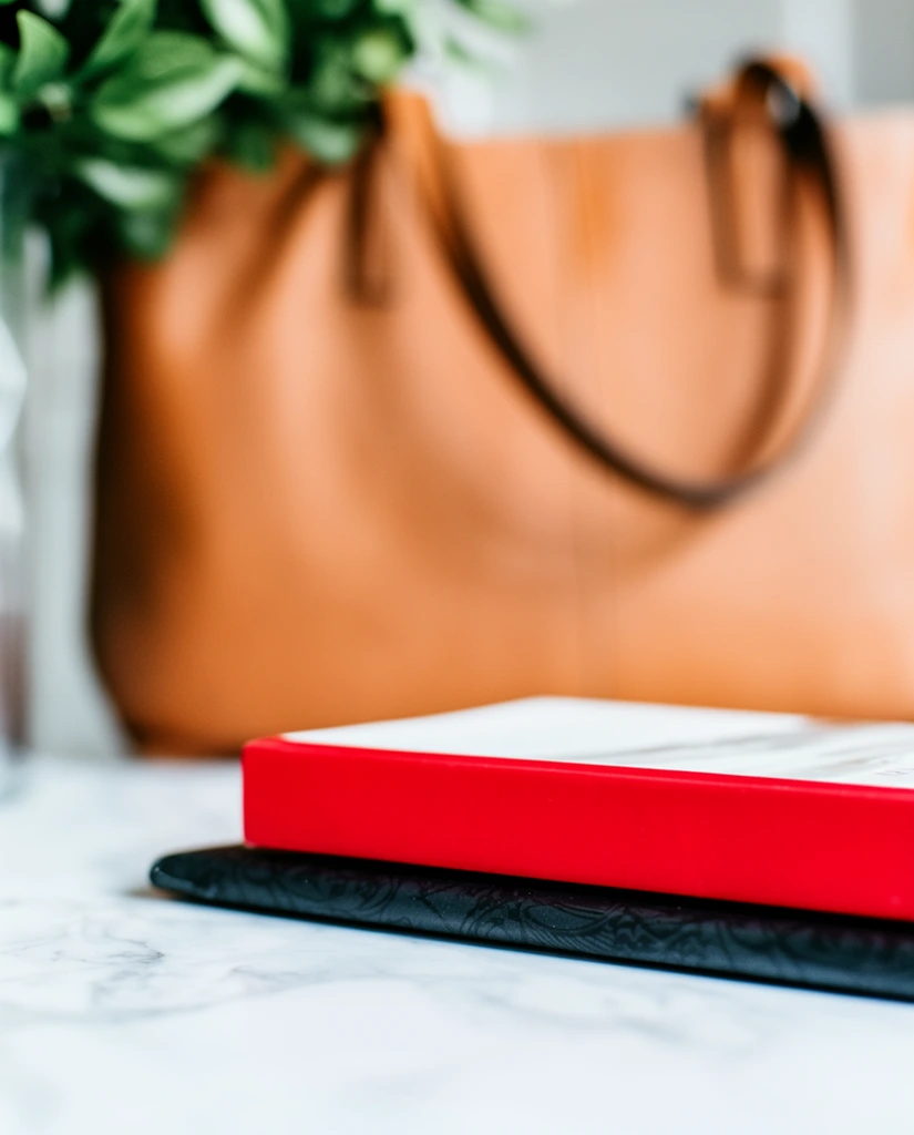 A close-up lifestyle photograph focusing on a closed book with a vibrant solid red spine resting horizontally atop a dark, subtly patterned notebook. The stacked items sit on a smooth white marble surface featuring faint grey veining. Behind the books, a warm, heavily blurred background displays a large tan leather tote bag with curved handles and a cluster of green leaves in the upper left corner. The composition utilizes a very shallow depth of field, keeping only the red spine and the dark notebook edge in sharp macro focus while rendering the background elements in soft bokeh. The lighting is bright and natural, creating a crisp contrast between the bold red accent and the neutral earth tones.