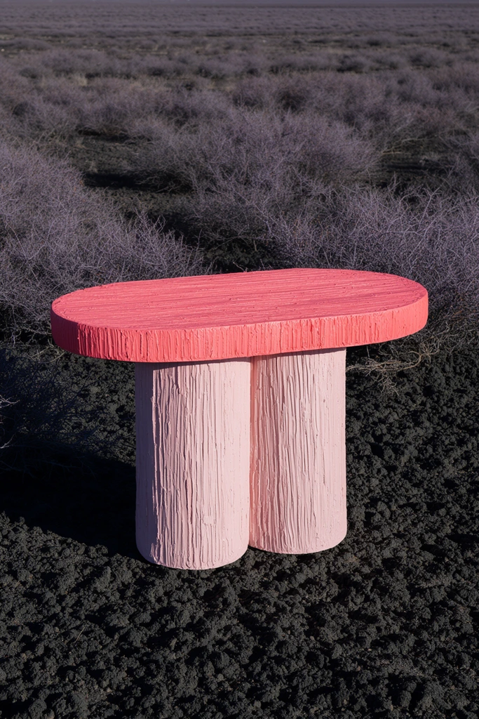 A brightly colored sculptural table rests in the center of a dark and arid landscape. The table features an oval top in deep salmon pink supported by a thick, double-cylindrical base in a lighter pastel blush. Its entire surface is heavily textured with deep, vertical striations resembling roughly carved wood or raked clay. The vibrant piece sits directly on clumpy, charcoal-colored earth, surrounded by low, leafless, purplish-grey scrub bushes stretching to a flat horizon. Direct sunlight angles from the right, casting a sharp, dark shadow to the left and sharply highlighting the deeply grooved texture of the furniture against the muted, barren environment.
