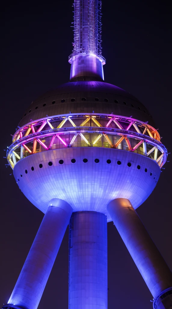 A giant spherical observation tower glows blue and purple against a dark night sky. Supported by massive diagonal struts, the metallic globe features an equatorial ring of geometric trusses illuminated by pink, yellow, and orange lights. A central vertical core extends upward. Low-angle telephoto night photography.