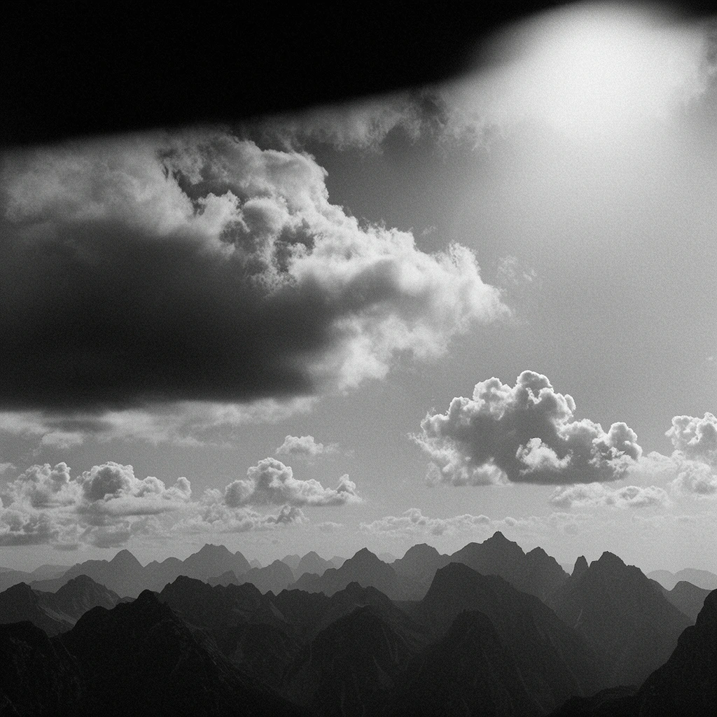 Black and white landscape photography, dark jagged mountain range silhouettes across the horizon, dramatic sky, heavy flat-bottomed storm cloud on the left, scattered cumulus clouds, high contrast, prominent film grain, heavy dark vignette at top edge, bright atmospheric light flare in upper right, moody atmospheric perspective
