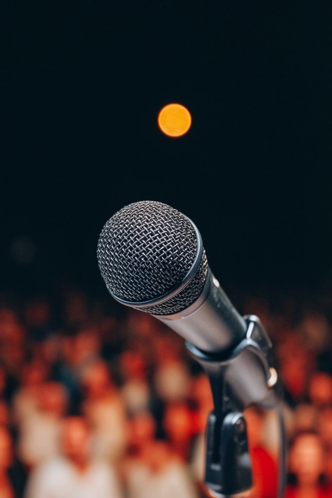 A dynamic close-up of a microphone standing on a stage, viewed from a low angle, dominating the foreground in sharp focus. The microphone has a dark grey wire mesh head and a matte black body, slightly angled towards the top left corner. Behind the microphone, the background is a vast, pitch-black space, punctuated by a single, perfectly round, glowing orange-yellow light in the upper center, resembling a distant moon or spotlight. In the lower half of the background, heavily blurred figures of a crowd are visible, appearing as indistinct blobs of red, orange, and white, creating a strong sense of depth and anticipation. The lighting is dramatic, focusing on the microphone, while the crowd and the single light source are softly blurred, enhancing the cinematic and atmospheric quality of the scene.