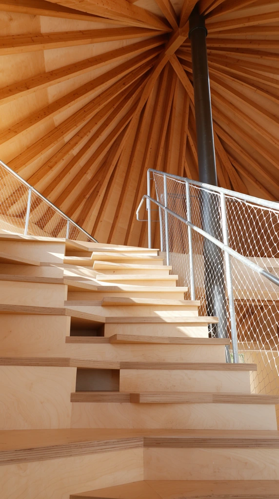 low angle view looking up modern alternating tread wooden stairs, light birch plywood construction, exposed wooden roof structure with radiating beams converging at the apex, dark cylindrical metal pipe running vertically, metal handrail with thin white mesh safety netting, warm natural sunlight, sharp cast shadows on wood surfaces, architectural photography, wide angle perspective, clean geometric design