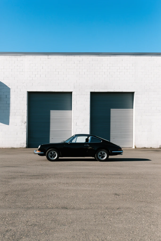 A vintage black sports coupe parks on an empty asphalt lot facing a white cinderblock industrial building with grey garage doors. Bright sunlight casts sharp shadows under a clear blue sky in this high-contrast minimalist photograph.