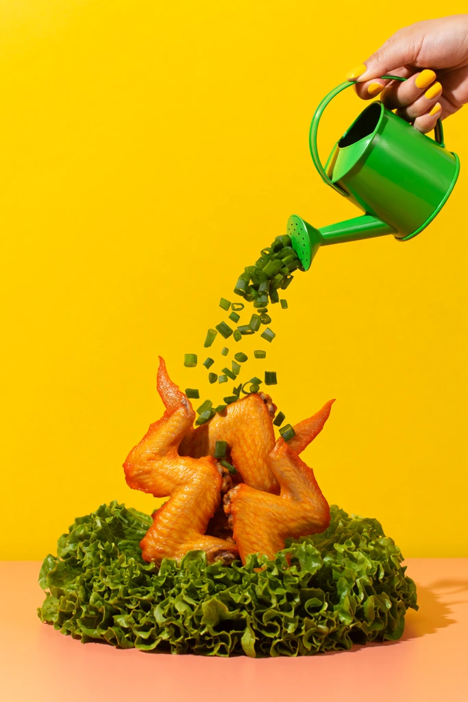 Surreal food photography, hand with yellow fingernails holding a bright green watering can pouring chopped green scallions, tight cluster of glossy golden-orange cooked chicken wings standing vertically below, wings planted in a dense bed of fresh curly green lettuce, solid vibrant yellow background, smooth peach-colored table surface, bold pop art aesthetic, bright studio lighting, highly saturated contrasting colors, playful composition
