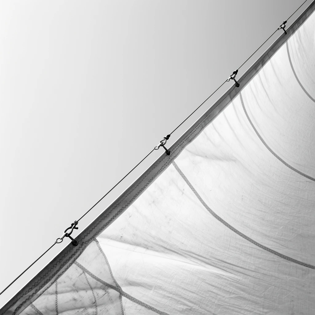 taut billowing boat sail catching wind, sharp diagonal composition from lower left to upper right, minimalist black and white photography, translucent fabric panels with distinct structural seams, reinforced canvas edges, metal shackles attached to parallel rigging lines, featureless light gray sky forming stark negative space, geometric abstraction, low angle perspective, crisp focus on textile texture and mechanical tension, high-key lighting