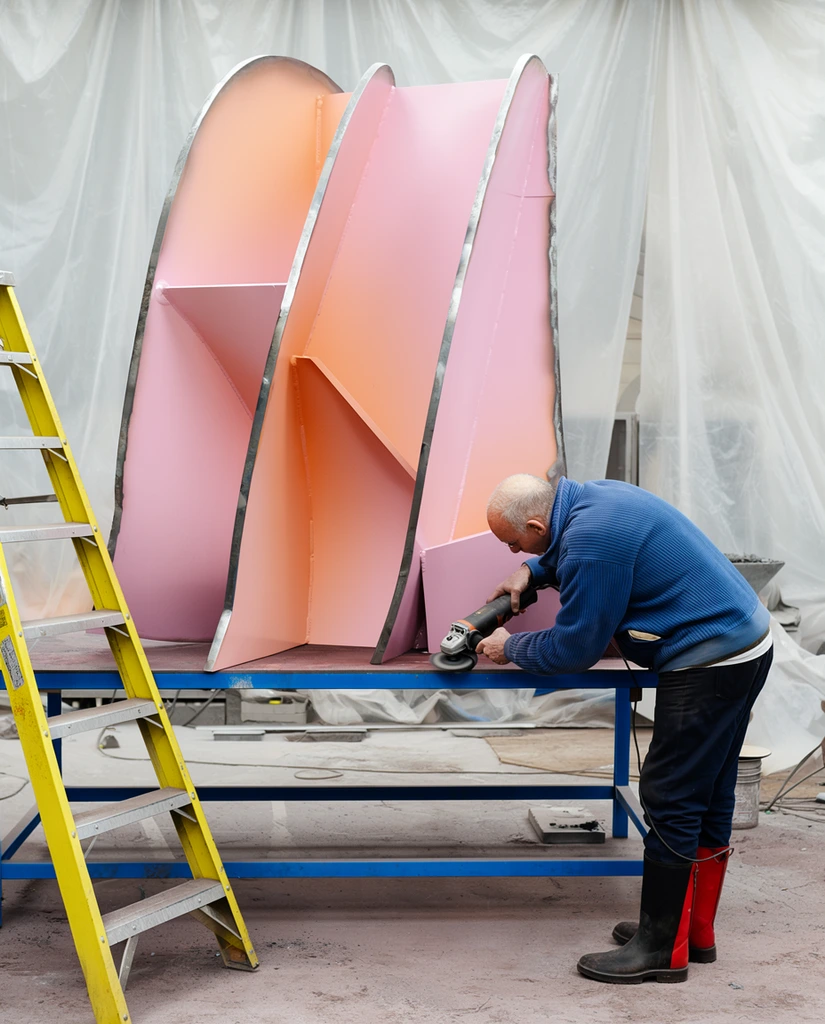 sculptor using an angle grinder on a massive pastel pink and peach metal sculpture, older man leaning forward on a blue work table, wearing a blue ribbed jacket and black boots with red side panels, abstract sculpture featuring intersecting curved and angular metal planes with raw grey metal edges, industrial workshop background draped with translucent white plastic sheeting, yellow stepladder on the left, dusty concrete floor, documentary photography, medium wide shot, natural lighting