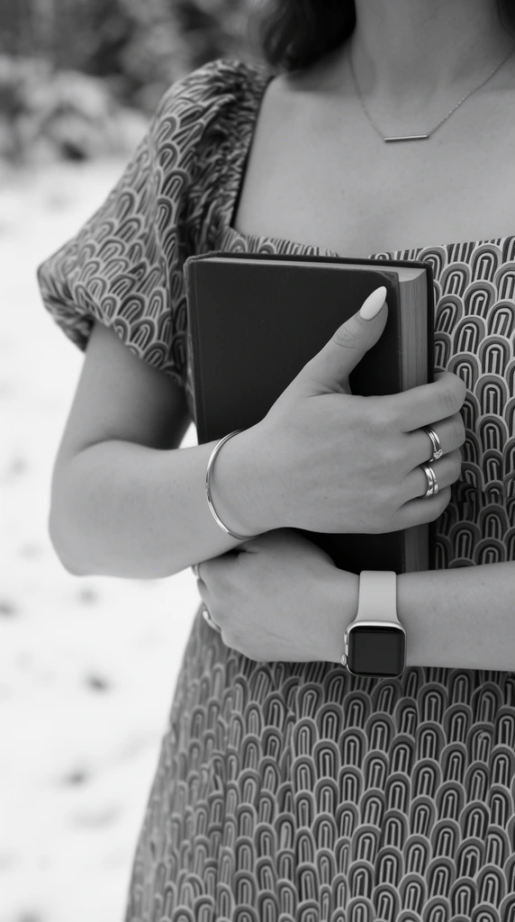 black and white mid-shot of a woman holding a closed dark book against her chest, wearing a patterned dress with short puff sleeves featuring repeating geometric arched shapes, delicate bar necklace, metallic rings, smooth manicured nails gripping the cover, thin metal bangle, modern smartwatch with a light solid strap, soft blurred snowy outdoor background, shallow depth of field, sharp focus on hands and book, smooth monochromatic tonal range