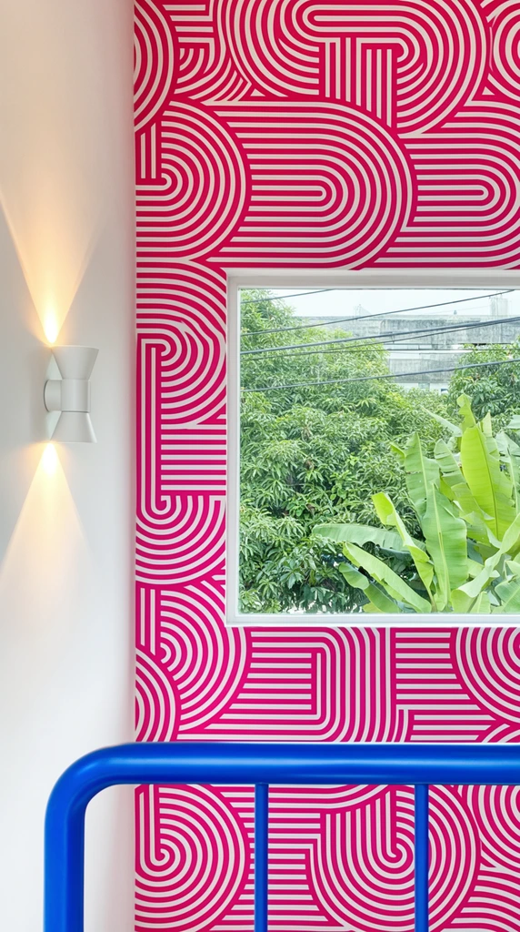 A modern interior corner defined by bold geometric patterns and vivid color contrasts. A dominant wall features striking magenta and white wallpaper with labyrinthine, interlocking semicircles and continuous parallel stripes. A rectangular window set within this vibrant pattern frames a lush exterior of dense green foliage, large banana leaves, overhead wire lines, and a distant grey building. To the left, a smooth white wall hosts a minimalist cylindrical wall sconce emitting warm, hourglass-shaped light beams upward and downward. In the immediate foreground, a bright cobalt blue tubular metal railing spans horizontally across the bottom edge.