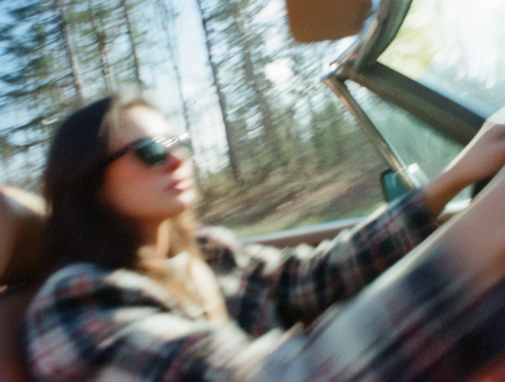 Heavy diagonal motion blur aggressively smears the form of a woman in sunglasses and a plaid jacket driving a convertible. The intense panning effect stretches the visual details into soft, rapid streaks of motion. Beyond the car's windshield, tall trees fill the background, their shapes elongated into continuous, blurred bands of color. The photograph features a raw, lo-fi snapshot aesthetic dominated by coarse film grain, high ISO noise, and a slightly muted, low-dynamic-range color palette. Bright, blown-out specular highlights streak across the frame, blending with the entirely soft, out-of-focus edges of the subjects and environment. The image captures a candid, high-speed moment rendered with the textural grit and slight color fringing characteristic of vintage, amateur film photography.