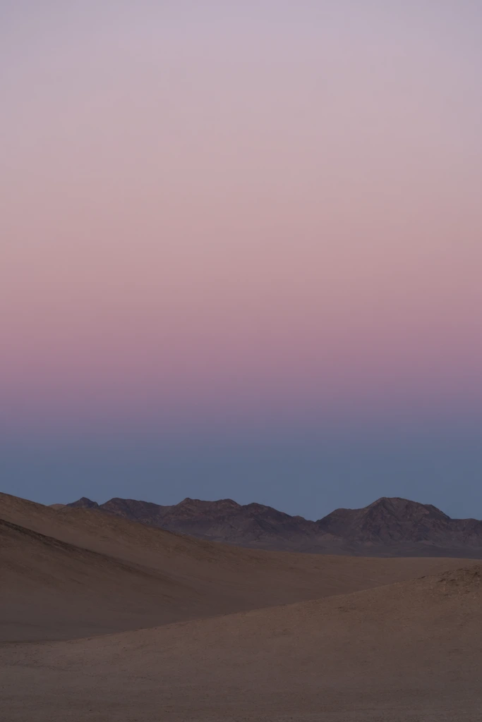 A minimalist barren landscape features undulating tan hills leading to distant, dark, jagged mountain peaks along the horizon. The upper two-thirds of the composition showcases a smooth twilight gradient sky, transitioning seamlessly from a deep dusky blue above the mountains into a soft lilac, and fading to a pale, dusty pink at the top. The wide horizontal framing emphasizes the vast open space, soft ethereal lighting, and distinct atmospheric color bands.