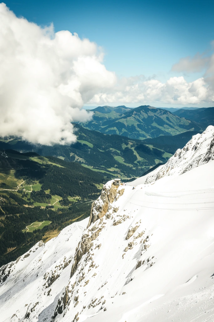 A high-altitude, wide-angle landscape photograph capturing a dramatic contrast between snow-covered alpine peaks and green valleys below. In the foreground on the right, a steep, rocky mountain slope is heavily blanketed in bright, sunlit white snow. A small, square structure sits precariously on a rocky outcrop protruding from this snowy slope, with tiny figures visible near it. Thin, dark cables stretch horizontally across the snowy expanse, suggesting a ski lift or cable car line. To the left, a deep valley unfolds, featuring dark green coniferous forests, lighter green patches of alpine meadows, and winding paths or roads. The background consists of distant, rolling mountain ridges in varying shades of blue and green under a bright blue sky. Massive, bright white cumulus clouds hover prominently on the left side, casting soft shadows on the peaks and valleys, adding a sense of immense scale and atmospheric depth to the mountainous terrain.