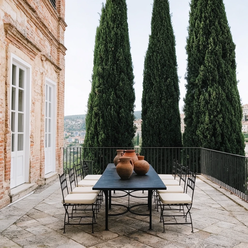 A paved stone patio centers on a long black metal dining table and matching chairs with cream cushions, displaying four rustic terracotta vessels. Three towering dark green cypress trees rise directly behind a metal railing against a pale sky, framing a distant hillside town. A weathered brick facade with white French doors flanks the left, captured in bright diffused daylight emphasizing sharp architectural lines.