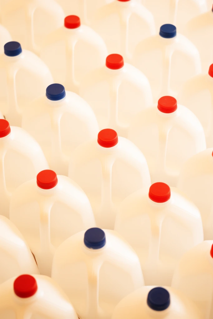 A dense overhead view of tightly packed white plastic gallon jugs filling the frame in a repeating pattern. The opaque containers feature molded handles and solid red or dark blue caps. Soft, diffused lighting casts a warm, high-key glow across the smooth curves. A shallow depth of field focuses sharply on the central diagonal row, softly blurring the surrounding jugs.