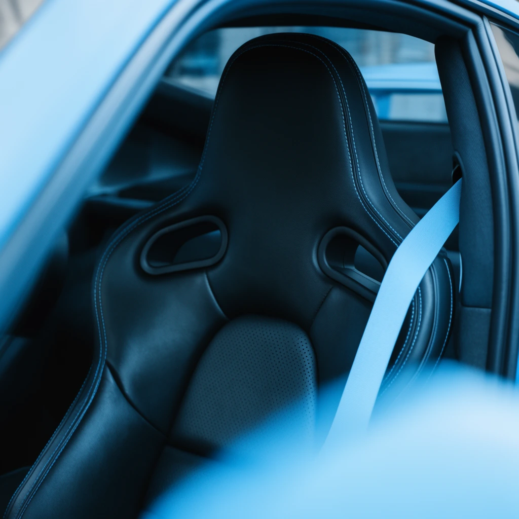 black leather racing seat, harness cutouts, contrast stitching, light blue seatbelt, blurred blue foreground frame, shallow depth of field, cool lighting