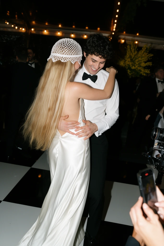 A couple dances closely on a glossy black and white checkerboard floor at a dimly lit event. The woman, viewed from behind, wears a sweeping backless white satin gown and a vintage-style white crocheted skullcap veil over her very long blonde hair. She embraces a smiling man with dark curly hair, dressed in a white dress shirt, black trousers, and a black bow tie. His hands rest on her waist as he looks down joyfully. The shadowy background features warm overhead string lights, blurred figures in dark suits, and a yellow floral arrangement. In the lower right foreground, hands holding a smartphone and a partial camera rig capture the scene. Harsh direct flash photography creates bright highlights on the crisp white garments and sharp shadows against the dark surroundings, emphasizing the candid, celebratory atmosphere.