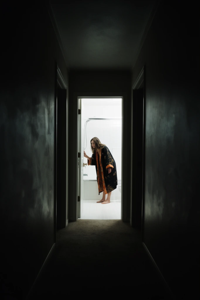 distant female figure framed within a brightly lit doorway at the end of a pitch-black narrow corridor, woman standing barefoot in a stark white tiled bathroom, wearing an oversized dark robe with orange and gold patterned lining, bending slightly forward, touching a glass shower door, symmetrical one-point perspective looking down a dark hallway, shadowy walls dominating the foreground, brilliant harsh white illumination spilling from the distant room, framing within a frame, cinematic wide shot, extreme high contrast, psychological thriller aesthetic, suspenseful atmosphere