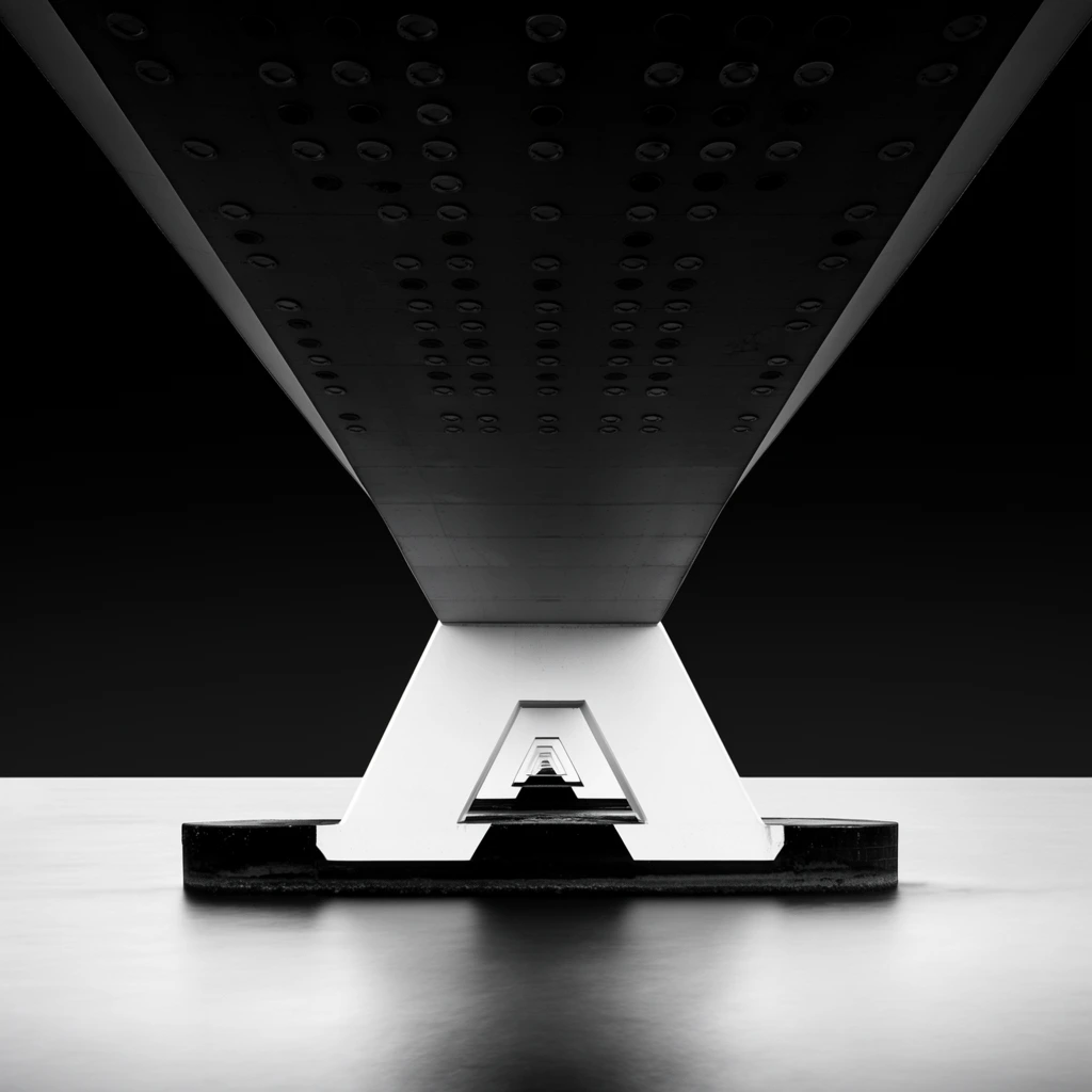 A highly symmetrical black and white architectural photograph capturing the underside of a modern bridge receding into a vanishing point. The stark composition is bisected by a crisp horizon line. A massive, dark bridge deck flares outward toward the top corners against a pitch-black sky, its surface detailed with neat rows of circular structural elements. At the exact center, a bright white trapezoidal concrete pier rests on smooth, light gray water rendered luminous by a long exposure. A hollow geometric cutout in the main pier frames a perfectly aligned sequence of receding identical supports. The minimalist, high-contrast framing emphasizes pure geometric form, one-point perspective, and surreal tranquility.
