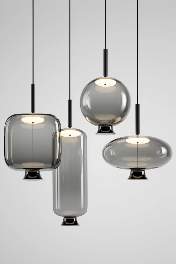 Four modern pendant lights hang against a seamless light grey background. Each features a transparent, smoky-grey glass shade in a distinct geometric form: a rounded square, a tall cylinder, a sphere, and a wide oval. Suspended by thin black cords with cylindrical fixtures, the lamps contain a glowing warm-white upper disc and a delicate central vertical wire dropping to a flared black bottom cap. Crisp studio reflections contour the pristine glass in this minimalist product showcase.
