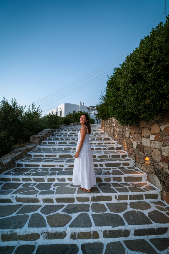 A smiling woman in a white maxi dress pauses on an ascending stone staircase, glancing back over her shoulder. The distinctive steps feature irregular grey flagstones heavily outlined in bright white paint. A rustic stone wall with warm embedded lights and lush green bushes borders the right side of the path. In the background, a partially obscured whitewashed building rests beneath thin power lines cutting diagonally across a clear, gradient blue twilight sky. The wide, low-angle perspective emphasizes the sweeping geometry of the stone walkway under soft, natural evening illumination.