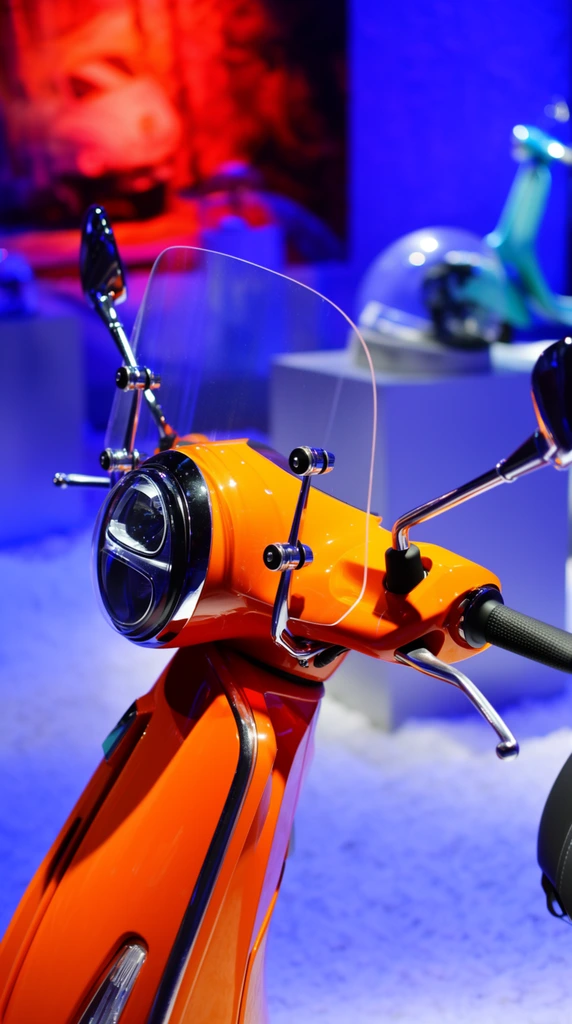 A glossy, bright orange retro-style motor scooter is captured in a close-up front profile, bathed in dramatic dual-tone lighting. The vehicle features a large round headlight with a split-reflector design, a clear acrylic windshield secured by polished silver brackets, and sleek chrome side mirrors. Textured black handgrips and curved metallic brake levers extend from the handlebars. The highly reflective front cowling catches the ambient light, contrasting with the textured white powdery surface below. A softly out-of-focus background reveals vibrant neon blue and fiery red lighting, illuminating blurred silhouettes of a display pedestal, a domed object, and a teal scooter. Shot with a shallow depth of field, the composition emphasizes the crisp details of the mechanical hardware and gleaming paint against the creamy bokeh of the brilliantly colored studio environment.