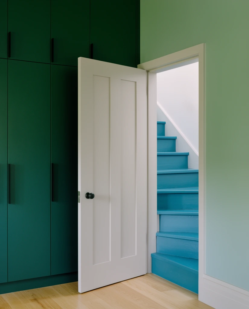 A modern interior features a striking color-blocked design with an open white paneled door revealing a bright teal staircase. Deep forest green floor-to-ceiling built-in cabinets with sleek black vertical handles dominate the left side. A soft sage green wall frames the right. The off-white door rests over light oak wood flooring, leading toward vibrant teal wooden steps illuminated by soft natural light against crisp white stairwell walls. The highly geometric architectural photography emphasizes clean vertical lines, minimalist aesthetics, and a rich analogous palette of varying green and blue hues.