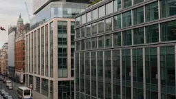 An office scene in 90s London. A superhero crashes through the building in an explosion of glass and paper, then morphs into an office worker mid-stride — one continuous handheld shot, shattering glass and settling paper in the mix.