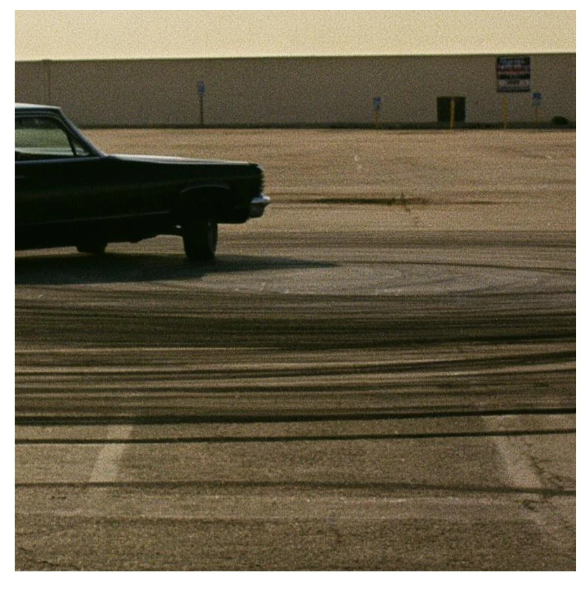 Cinematic still of a black vintage sedan parked in an empty concrete lot at dusk, muted film-grain color grade, wide anamorphic framing, long soft shadows.