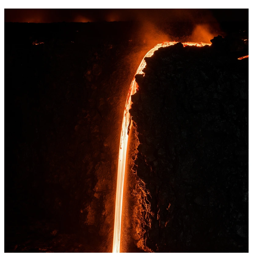 Molten lava pouring over a dark volcanic ledge at night, a glowing orange river against black basalt, long-exposure heat glow, dramatic high-contrast lighting.