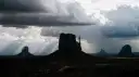 Dramatic desert landscape with towering buttes silhouetted against a stormy sky. Dark rain clouds loom overhead as sunbeams break through gaps, casting striking rays of light across the rugged terrain and highlighting the iconic rock formations