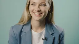 Happy, playful 26-year old caucasian woman, studio shot for interview, from B-camera footage. Wearing a blue blazer and white t-shirt. Making eye contact with viewer, then looking slightly off screen, in silence. Mouth closed. Gentle smile. Mint green background. Stylized lighting. No dialogue. No technical equipment in shot.