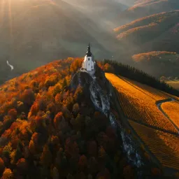 A stark white church with a dark spire crowns a steep hilltop surrounded by a vibrant autumn landscape. The bright architecture contrasts sharply against rolling forests of fiery orange, gold, and deep green foliage. Terraced vineyards sweep down the sunlit right slope. Captured from an elevated aerial perspective, dramatic low-angle golden light illuminates the crest, casting the background valleys into deep, misty shadow.
